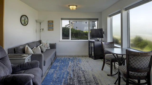 Cozy living room with a gray sofa, blue patterned rug, wall clock, floor lamp, TV on a cabinet, and a dining table by large windows overlooking a sunny outdoor view.