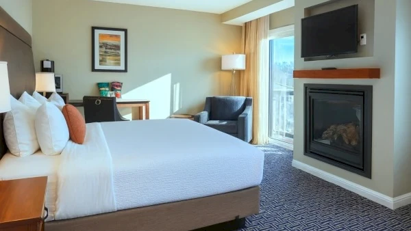 A hotel room with a king bed, nightstands, a desk, a chair, a fireplace, a mounted TV, a balcony door, and neutral decor ending with a period.