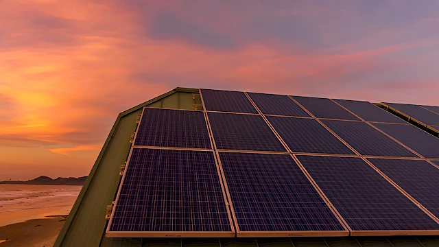 A row of solar panels on a sloped roof by a beach at sunset, casting a warm orange glow across the sky and water.