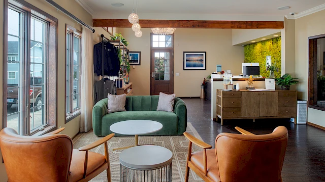 A modern, cozy lobby with a green sofa, two brown leather chairs, round white coffee tables, and a white circular side table; large windows, a reception desk, and indoor plants.