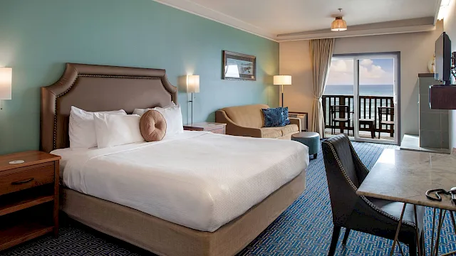 A hotel room with a king bed, teal walls, nightstands, lamps, a sofa, desk, and a balcony overlooking the ocean.